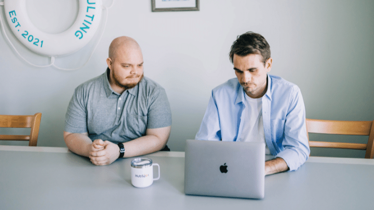 two men sitting at a table with a laptop and mug
